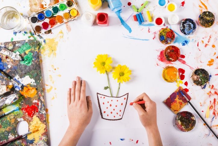 Person painting a flower pot on table full of art supplies.